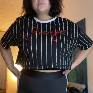 Croptop med text - Svart, lite boxig croptop med vita ränder rött broderi med texten ”Stranger”. Köpt på New Yorker. Använd ca 5-7 gånger. Haft ett hål men lagat det (bild 3) och tycker inte det stör då det är på baksidan av axeln/nära axelsömmen. Har tvättats efter lagning och den håller. 💌Frakten är baserad på frimärken. Önskas postnords egna paket löser vi det.🎈