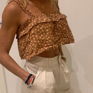 Brunt croppat linne - Gulligt brunt linne med blommor och volanger. Använd 1 gång och är i sitt orginala skick (orginal pris 170kr)