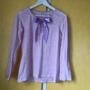 En skön långärmad tröja i pastell rosa färg. Den har en lila rosett och spets runt händer och krage