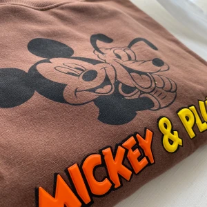 Mickey & Pluto hoodie  - Brun hoodie med tryck av Musse och Pluto. Superskön ock armarna är så fina då de är lite kortare.  