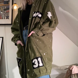 Militärgrön trenchcoat  - Militärgrön trenchcoat med svart och vita fläckar samt påsydda tryck. Med snörning i huvan och längst ner. Knälång och svincool men har inte använt på länge. I mycket gott skick! Från forever 21 i storlek M, Köparen står för frakt! 