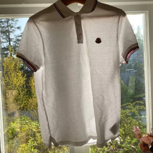 Vit piké t-shirt från Moncler/byter endast - Vit piké t-shirt från Moncler med klassisk krage och knappar framtill. Ärmar och krage har röd och marinblå randig detalj. Moncler-logga på bröstet. Perfekt för en stilren och sportig look.