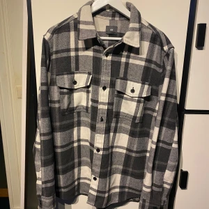 Svartvit rutig skjorta från H&M - Säljer en svartvit rutig skjorta från H&M i storlek L. Skjortan har klassisk krage, knäppning framtill och två bröstfickor med lock och knapp. Perfekt för lager-på-lager och en avslappnad stil.
