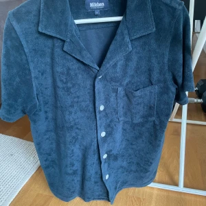 Mörkblå frottéskjorta från Nikben - Säljer en mörkblå kortärmad skjorta från Nikben i mjuk frotté. Skjortan har klassisk krage, knappar framtill och en bröstficka. Perfekt för en avslappnad och cool stil. Köpt för 1200