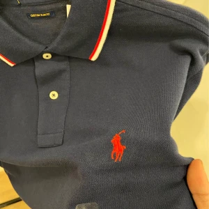 Blå pikétröja från Ralph Lauren - Säljer en blå pikétröja från Ralph Lauren med röd logga broderad på bröstet. Tröjan har vit och röd kant på kragen och ärmarna samt två knappar framtill. Modellen är custom slim fit och passar perfekt för en stilren look.