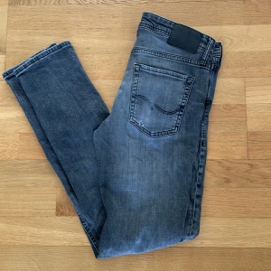 Grå slim fit jeans - Säljer ett par grå slim fit jeans från Jack & Jones med klassisk femficksdesign och diskreta slitningar på vänster lår. Jeansen har normal midja och stängs med knappar. Perfekta för en avslappnad stil.