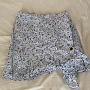 Ljusblå blommig kjol med volang - Söt ljusblå kjol  som är använd fåtal gånger. Kjolen har volang längs nederkanten och en knappdetalj på sidan. Perfekt för en somrig look och har en lätt och luftig känsla.