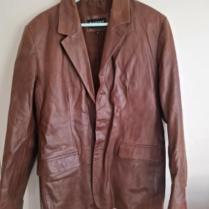 Brun skinnkavaj från HOLLIES - Säljer en brun kavaj i äkta skinn från HOLLIES. Klassisk modell med krage, knäppning framtill och två fickor med lock. Perfekt för dig som vill ha en stilren och tidlös jacka i garderoben.