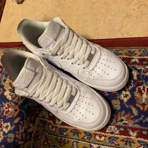 Vita Nike Air Force 1 sneakers - Klassiska vita Nike Air Force 1 sneakers med perforerad tå och platt sula. Skorna har snörning och är tillverkade i skinn med diskreta Nike-loggor på sidan och tungan. Perfekta för en clean och stilren look.