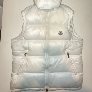 Vit dunväst från Moncler - Säljer en snygg vit dunväst från Moncler med dragkedja och två fickor framtill. Västen har en hög krage och klassisk Moncler-logga på bröstet. Perfekt för lager-på-lager och stilren look. Ingen Frakt❌ 