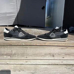 Svarta sneakers från Philippe Model - Säljer ett par svarta Philippe Model sneakers med vita detaljer och svart snörning. Skorna har en klassisk låg profil, rund tå och platt sula. Materialet är en mix av mocka och syntet, med märkets logga på sidan. Skrona är i nästan nyskick knappt använda, medföljer dock inte låda eller sulor 