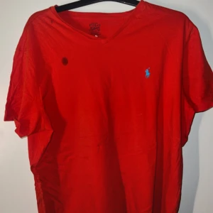 Ralph Lauren tröja  - Säljer en röd t-shirt från Polo Ralph Lauren med blå broderad logga på bröstet och v-ringad hals. T-shirten har korta ärmar och är i mjuk bomull. Perfekt för dig som gillar klassisk stil med en färgklick. Dock en fläck 