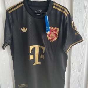 Svart Bayern München fotbollströja Musiala 42 - Säljer en svart Bayern München fotbollströja från Adidas med guldiga detaljer, klubbmärke och Musiala 42 på ryggen. Tröjan har korta ärmar, rund hals och klassiska Adidas-ränder på axlarna. Perfekt för fans av Musiala och Bayern.