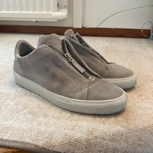 Grå sneakers från Axel Arigato med dragkedja - Säljer ett par stilrena grå sneakers från Axel Arigato i mocka med dragkedja framtill istället för klassisk snörning. Skorna har vit platt sula och diskret logga i guld på sidan. Perfekta för en clean och modern look. Avvända max 10 gånger alltså väldigt bra skick 