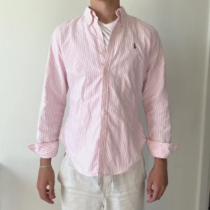 Rosa och vit randig skjorta från Ralph Lauren - Säljer en klassisk rosa och vit randig skjorta från Ralph Lauren med button-down krage och broderad logga på bröstet. Skjortan har långa ärmar och knäppning framtill. Perfekt för en stilren och fräsch look.