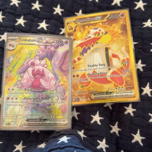 Pokémonkort Tinkaton ex och Skeledirge ex - Två Pokémonkort: Tinkaton ex med holografisk yta och 300 HP, samt Skeledirge ex med 340 HP och guldfärgad design. Båda korten är officiella Pokémonkort på engelska och har färgstarka illustrationer. Perfekt för samlare eller Pokémonfans.