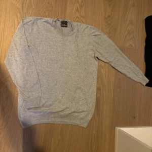 Ljusgrå tröja från Oscar Jacobson - Säljer en stilren ljusgrå tröja från Oscar Jacobson i storlek XL. Tröjan har rund halsringning och långa ärmar, perfekt för lager-på-lager. Mjuk och bekväm, passar till många olika outfits.
