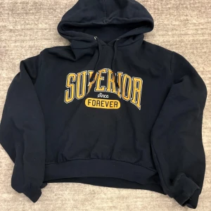 Mörkblå hoodie med tryck från Superior - Säljer en mörkblå hoodie från Superior med gul text 'Superior since Forever' på bröstet. Tröjan har huva med dragsko och ribbade muddar vid ärmslut och nederkant. Perfekt för en avslappnad stil.