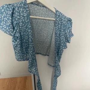 Blommig blå omlottopp från SHEIN - Säljer en söt blå omlottopp från SHEIN med vitt blommönster. Toppen har korta puffärmar och knytband i midjan. Perfekt för vår och sommar!