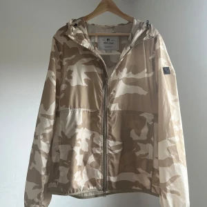 Camo windbreaker från Woolrich🐑 - Säljer en beige och vit camouflage vindjacka från Woolrich med huva och dragkedja framtill. Jackan har en avslappnad passform och är perfekt för blåsiga dagar. Det är en äldre model av Woolrich väldigt unika vindjackor. Stilsäker detalj med logotyp på ärmen väldigt bra skick vid minsta lilla fundering är det bara att höra av sig✌️