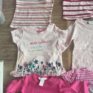 9-pack samling långärmade tröjor för barn - 9 stycken långärmade tröjor i olika färger och mönster, bland annat randigt, enfärgat och med blommor. Färgerna går i rosa, vitt och rött. Några tröjor har tryck som 'mariposa' och 'MEXX'. Perfekt för dig som gillar mjuka och bekväma plagg.