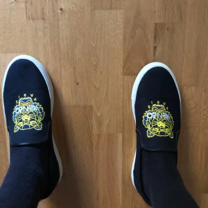 Svarta slip-on sneakers med tigerbrodyr från Kenzo - Svarta slip-on sneakers från Kenzo med en färgstark gul och blå tigerbrodyr på ovandelen. Skorna har vit platt sula och är tillverkade i textil med Kenzo Paris-logga på insidan. Perfekta för en cool och avslappnad stil.
