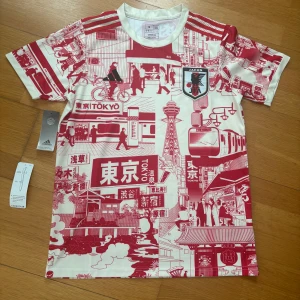 Japan Tokyo special edition fotbollströja från Adidas - Unik fotbollströja från Adidas med Tokyo-inspirerat rött och vitt mönster. Tröjan har korta ärmar, rund hals och Japan-emblem på bröstet. Perfekt för dig som gillar japansk kultur och fotboll.