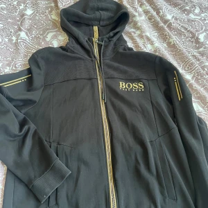 Svart hoodiejacka från Hugo Boss - Svart hoodiejacka från Hugo Boss med gulddetaljer och dragkedja framtill. Jackan har huva med snörning och logga på bröstet samt ärmar med dekorativa ränder. Perfekt för en sportig och stilren look.