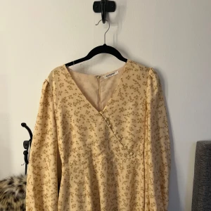 Blommig midsommarklänning från Glamorous - Söt ljusgul/beige klänning från Glamorous med små blommor i ett diskret och elegant mönster. Klänningen har omlott-skärning framtill, lång ärm och dragkedja i ryggen. Perfekt för vår och sommar! Säljes då den inte kommer till användning, skulle även kunna passa M. Nypris 1200kr