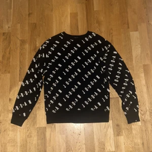 Svart Jordan sweatshirt med logotypmönster - Säljer en svart sweatshirt från Jordan med vitt logotypmönster över hela tröjan. Tröjan har rund halsringning och långa ärmar. Perfekt för dig som gillar sportig och stilren streetwear.