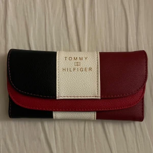 Plånbok i rött, vitt och svart - Säljer en snygg plånbok med klassiska färgblock i rött, vitt och svart. Plånboken har guldfärgad logga framtill och är tillverkad i syntetmaterial. Perfekt accessoar för att hålla ordning på kort och kontanter.