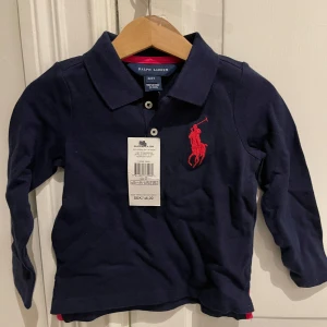 Ny Mörkblå pikétröja från Ralph Lauren - Säljer en ny klassisk mörkblå pikétröja från Ralph Lauren med stor logga broderad på bröstet. Tröjan har krage, knappar framtill och långa ärmar. Perfekt för en stilren och sportig look.