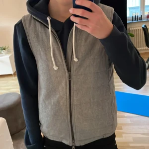 Ljusgrå quiltad väst från Oscar Jacobson - Säljer en stilren ljusgrå quiltad väst från Oscar Jacobson. Västen har dragkedja framtill, två sidfickor och diskreta knappar nedtill. Perfekt att bära över en hoodie eller skjorta för en snygg lager-på-lager-look.
