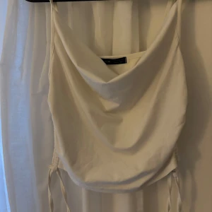 Vit croppad linnetopp med draperad hals - Säljer en vit croppad linnetopp med draperad halsringning och smala axelband. Toppen har justerbar snörning i sidorna för en snygg passform. Perfekt för varma dagar och enkel att styla till olika looks.