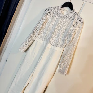 Vit byxdress med spetsdetaljer - Säljer en elegant vit byxdress med långärmad överdel i spets och hög midja. Byxorna är vida och raka, vilket ger en stilren look. Perfekt för dig som vill sticka ut med en unik och trendig outfit.