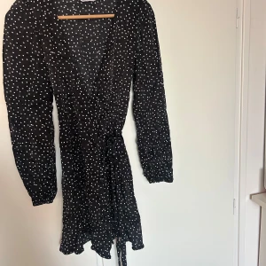 Svart omlottklänning med vita prickar från & Other Stories - Säljer en svart omlottklänning från & Other Stories med vita prickar. Klänningen har lång ärm, knyt i midjan och v-ringad hals. Perfekt för dig som gillar klassiska mönster och en feminin siluett.