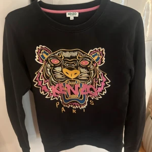 Svart sweatshirt med tigerbroderi från Kenzo - Cool svart sweatshirt från Kenzo med ikoniskt tigerbroderi i flera färger och texten 'Kenzo Paris' framtill. Tröjan har rund halsringning och långa ärmar. Perfekt statement-plagg för dig som gillar streetstyle. Pris kan absolut diskuteras!