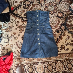 Jeansklänning med knappar - Säljer en snygg axelbandslös jeansklänning med knappar framtill och fickor. Klänningen har en figurnära passform upptill och lätt utställd kjol. Perfekt för dig som gillar denim och vill ha en cool look.