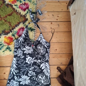 Svartvit blommig halterneckklänning - Säljer en svartvit kortklänning med blommigt mönster och halterneck med knyt i nacken. Klänningen har en tight passform och är ärmlös, perfekt för varma dagar.