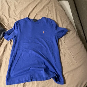 Blå t-shirt från Polo Ralph Lauren - Säljer en blå t-shirt från Polo Ralph Lauren med klassisk passform och korta ärmar. T-shirten har en liten broderad logga i orange på bröstet. Storlek S/M