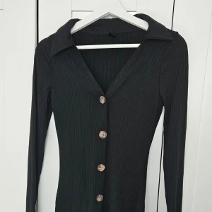 Svart ribbad långärmad klänning med knappar - Säljer en svart långärmad klänning med ribbad struktur och stora knappar framtill. Klänningen har en djup v-ringning och klassisk krage, vilket ger en stilren och modern look.