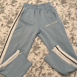 Ljusblåa träningsbyxor från Palm Angels - Säljer ett par ljusblåa träningsbyxor från Palm Angels med vita och svarta ränder längs sidorna. Byxorna har resår och snörning i midjan, dragkedjor vid bensluten och två fickor fram. Palm Angels-logga tryckt på låret.