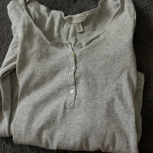 Ljusgrå ribbad långärmad tröja med knappar - Säljer en ljusgrå långärmad tröja i ribbat material med knappdetaljer framtill. Tröjan har en enkel och stilren design som passar till mycket. Perfekt för lager-på-lager eller som den är.