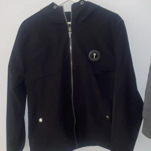 Svart windbreaker  från Trapstar - Säljer en svart vindjacka från Trapstar med huva och dragkedja framtill. Jackan har Trapstar-logga på bröstet och broderad text på ryggen. Snygga metalldetaljer med T på dragkedjan och fickor med tryckknappar ock in graverat T på de. Snören till jackan tillkommer vid önskemål!