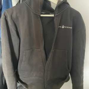 Svart hoodie från SAIL RACING - Svart hoodie med dragkedja från SAIL RACING. Tröjan har två stora fickor framtill och en liten logga på bröstet. Perfekt för en avslappnad stil och enkel att matcha med det mesta.