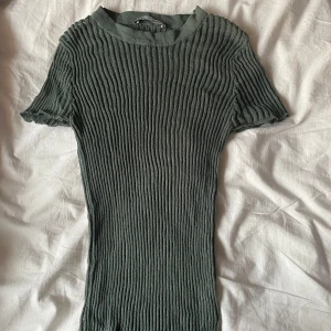 Grön ribbad topp från Zara - Säljer en grön ribbad topp från Zara med korta ärmar och rund halsringning. Klänningen har en tight passform och är tillverkad i ett stretchigt material som ger en snygg siluett. Perfekt för dig som gillar stilrena och bekväma plagg.