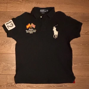 Svart pikétröja med tryck från Polo Ralph Lauren - Svart pikétröja med stor vit polospelare på bröstet, broderade flaggor och text på vänster sida samt stort nummertryck på ärmen och ryggen. Gula bokstäver 'SPAIN' på ryggen. Klassisk krage och knappar framtill.