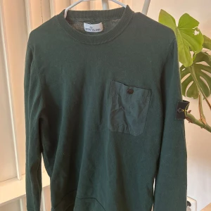 Mörkgrön tröja från Stone Island - Säljer en mörkgrön långärmad tröja från Stone Island med rund halsringning och en bröstficka med knapp. Klassisk logotyp-patch på ärmen. Tröjan är i mjukt material och har ribbade muddar vid hals, ärmslut och nederkant.