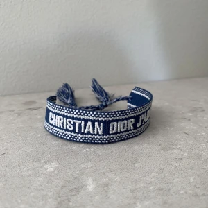 Dior armband - Säljer ett blått tygarmband från Christian Dior med texten 'Christian Dior J'Adior' i vitt. Armbandet har justerbar knytning med tofsar i ändarna och en snygg kontrastkant. Bara att höra av sig vid minsta funderingar eller frågor!🙌🌟