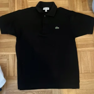 Svart pikétröja från Lacoste - Aldrig använt Helt ny köpte för 699kr Klassisk svart pikétröja från Lacoste med den ikoniska gröna krokodilloggan broderad på bröstet. Tröjan har korta ärmar, krage och knappar framtill. Perfekt för en stilren och sportig look.